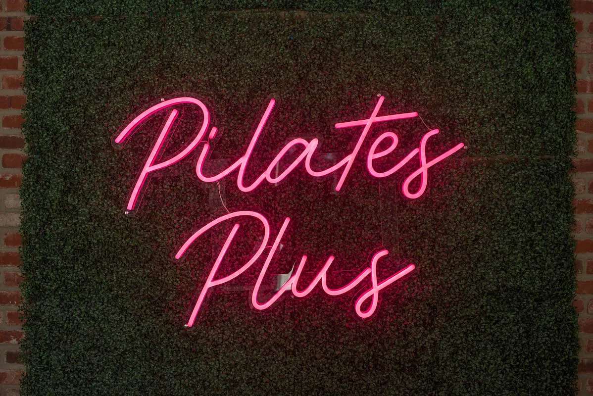 Pink neon Pilates Plus sign on a greenery wall